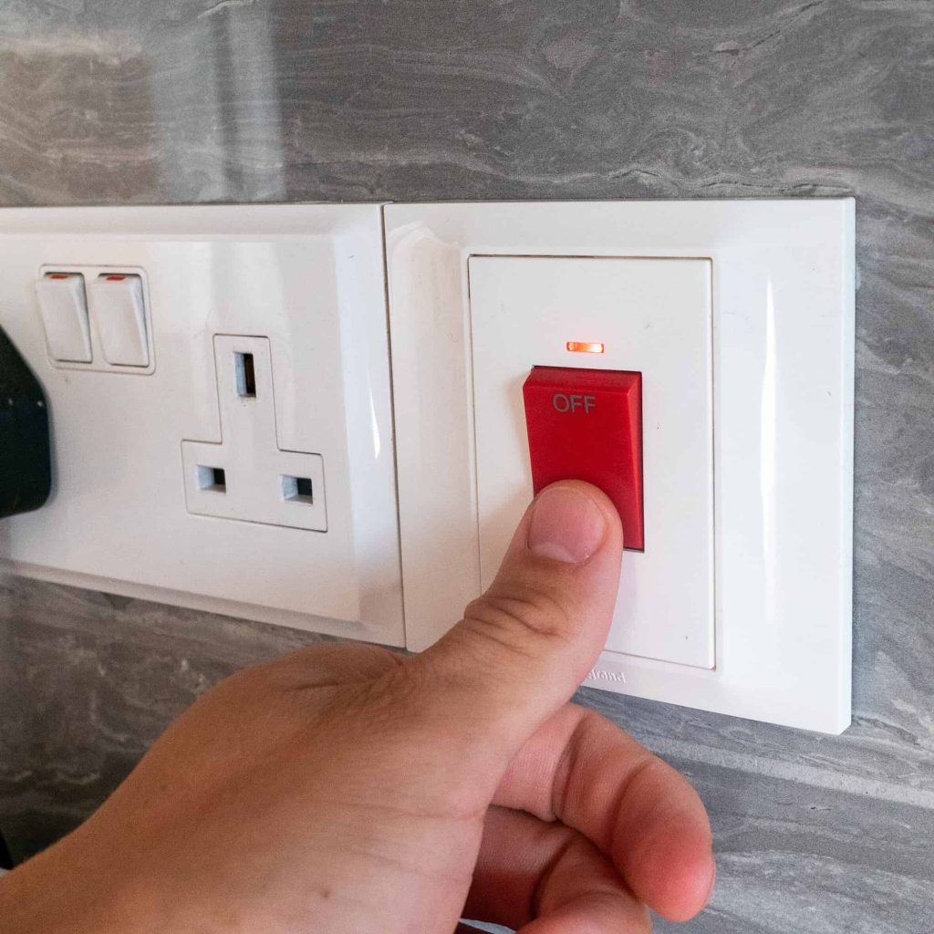 A hand is pressing the red "OFF" switch on a wall-mounted electrical outlet with a grey marbled background. A small red indicator light above the switch is illuminated.