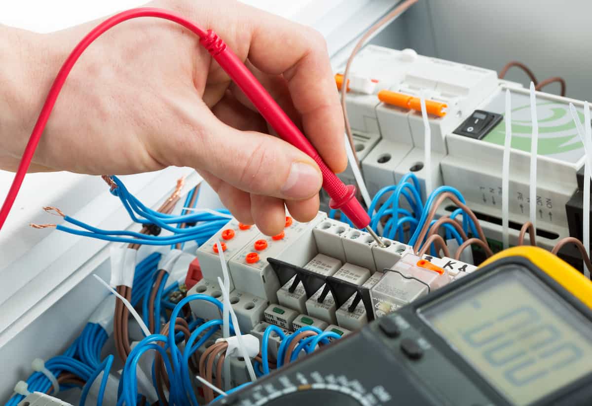 A person uses a red multimeter probe to test connections on an electrical control panel filled with blue and brown wires, components, and a digital multimeter display.